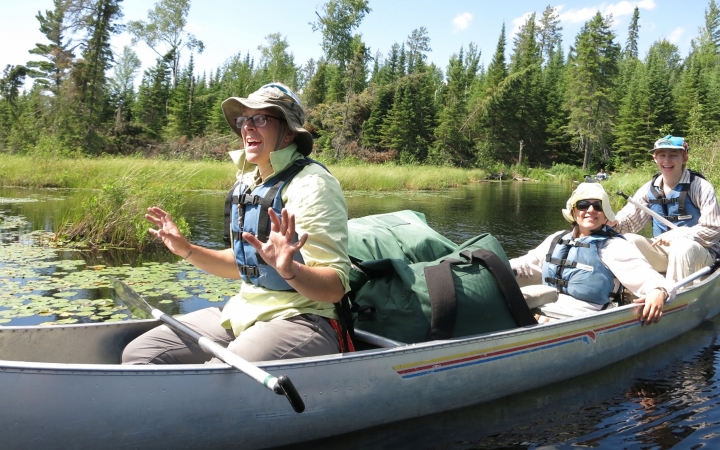 Three people sit in a canoe on calm water. Behind the canoe, lily pads rest on the water, with grass and a line of evergreen trees in the background.
